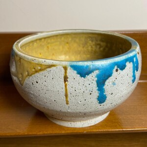 May include: A speckled ceramic bowl with a white exterior, featuring drips of yellow and blue glaze. The interior is yellow, and the bowl sits on a small, round base. The bowl is on a wooden surface.