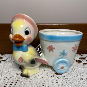 May include: A vintage ceramic planter shaped like a duck pulling a cart. The duck has a yellow body, pink hat, blue bow tie, and orange beak. The cart is white with blue trim and floral accents. A charming decorative piece.