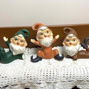May include: Three ceramic elf figurines with painted faces, white beards, and pointed hats. The left elf wears a green outfit, the center elf wears an orange outfit, and the right elf wears a brown outfit. Each elf has black pointed shoes.