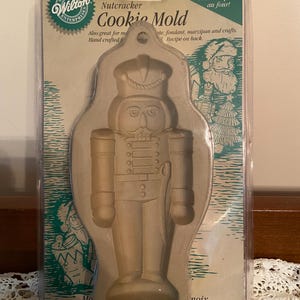 May include: A beige plastic Nutcracker Cookie Mold by Wilton Enterprises. The mold is in a clear plastic package and features a detailed design of a nutcracker soldier. The packaging includes text that reads "Ovenproof!" and "Cookie Mold."