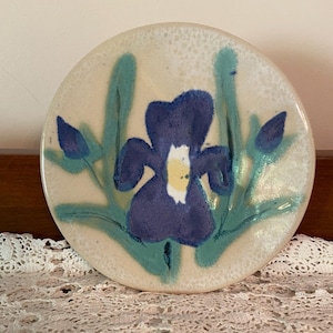 May include: Round ceramic plate with a hand-painted iris design. The iris is a deep blue with a yellow center, surrounded by green leaves and buds. The plate has a speckled, off-white background.