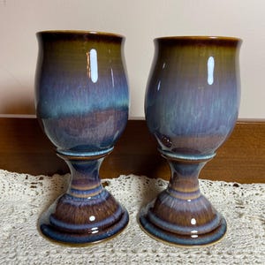 May include: Two ceramic goblets featuring a glossy, multi-toned glaze that transitions from a deep brown at the rim and base to a vibrant blend of purple and blue in the center. Each vessel has a rounded bowl, a slender stem, and a wide base.