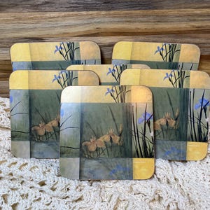 May include: Set of six square coasters with a floral design. The coasters feature a watercolor-style painting of irises in shades of yellow, blue, and green. The top border is yellow and blue. The bottom right corner of each coaster has the word "Pinefields".