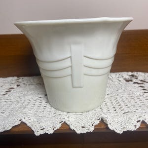 May include: A white ceramic vase with a flared top and a series of horizontal bands around the body. A vertical rectangular accent is centered on the front. The vase is sitting on a white lace doily.