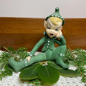 May include: A ceramic elf figurine with a matte green finish, wearing a pointed hat with white polka dots. The elf has blonde hair, painted facial features, and is posed in a seated position with one hand resting against its cheek.