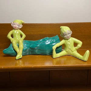 May include: Two ceramic elf figurines in light yellow outfits with pointed hats and ears. One elf sits atop a textured, dark teal hollow log planter, while the other elf sits on the surface with legs extended, both featuring painted facial details.