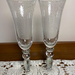 May include: Two clear champagne flutes with a bubble design. The flutes have a flared top, a stem with three spherical accents, and a round base. The glasses are made of clear glass and are displayed on a white lace tablecloth.