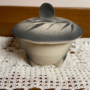 May include: A ceramic lidded jar with a speckled beige body and a dark gray lid. The lid features a leaf design and a small, textured handle. The jar sits on three small feet and is displayed on a white, crocheted doily.