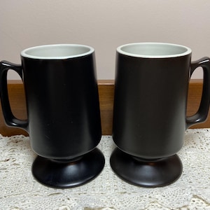 May include: Two matching mugs with a matte black exterior finish and a smooth white interior. Each mug features a tall, cylindrical body, a sturdy side handle, and a wide, circular pedestal base for stability. The design is simple and uniform.