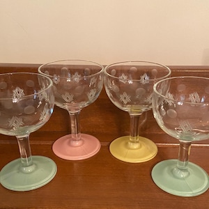 May include: Four clear champagne glasses with etched dot and geometric designs. Each glass has a colored base: green, pink, yellow, and green. The glasses are arranged on a dark wooden surface.