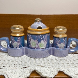 May include: A vintage ceramic salt and pepper set with a sugar bowl. The set features a blue base with white floral designs and gold accents. The tray and handles are a light purple color. The sugar bowl has a lid with a small spoon opening.