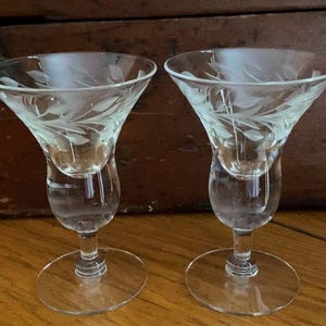 May include: Two clear glass cordial glasses with etched leaf designs. The glasses have a flared bowl, a bulbous midsection, and a circular base. The stemware is suitable for serving after-dinner drinks.