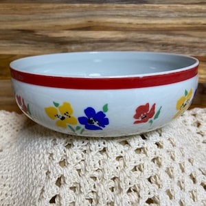 May include: White ceramic bowl with a red rim and colorful floral designs. The flowers are yellow, blue, and red, adding a cheerful touch. The bowl sits on a cream-colored, open-weave fabric.