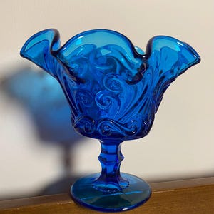 May include: A vibrant blue glass pedestal bowl with a ruffled rim. The bowl features an intricate swirl pattern and sits on a detailed stem and circular base. The glass has a glossy finish.