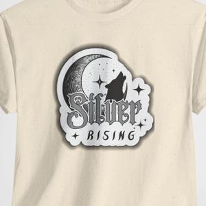 May include: Cream-colored t-shirt featuring a graphic design. The design includes a crescent moon, a howling wolf silhouette, and the words "Silver Rising" in a decorative font. The design is in shades of gray and white.