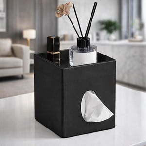 Op de afbeelding: Een zwarte vierkante tissuebox met een bovenste plank met een lippenstift, een glazen diffuser met zwarte stokjes en een decoratieve bloem. Witte tissues zijn zichtbaar door de ovale opening. De tissuebox staat op een wit oppervlak.
