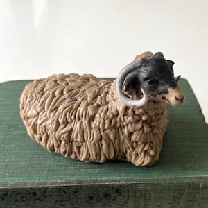 Puede incluir: Figura detallada de un carnero, con cuerpo beige texturizado y cabeza negra. El carnero tiene cuernos curvados y descansa sobre una superficie verde. Objeto decorativo.