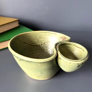Op de afbeelding: Een keramische kom met een gespikkelde lichtgroene glazuurlaag, voorzien van een groot hoofdcompartiment en een kleiner aangehecht zijcompartiment. De binnenzijde van de grote kom toont zichtbare horizontale ribbels en de buitenzijde is glad.