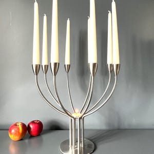 Op de afbeelding: Een zilverkleurige metalen kandelaar met een ronde voet en zeven gebogen armen die lange, slanke, gebroken witte kaarsen vasthouden. Een klein, brandend waxinelichtje staat in het midden van de voet. Twee rode appels liggen links.