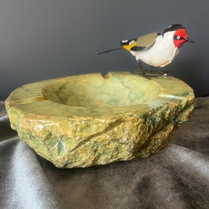 Op de afbeelding: Een ronde, groene, getextureerde stenen asbak met een ruwe, natuurlijke buitenkant en een gladde, gepolijste binnenkant. Een kleine, decoratieve metalen vogelbeeldje met een rood gezicht, wit lichaam en zwart-gele vleugeldetails zit op de rand.