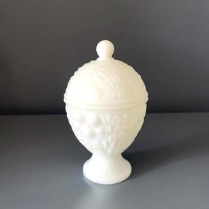 Può includere: Una bomboniera bianca con coperchio e base a piedistallo. Il piatto è realizzato in vetro opaco e presenta motivi floreali e a stella in rilievo. Il coperchio ha un piccolo pomello rotondo. Un classico elemento d'arredo.