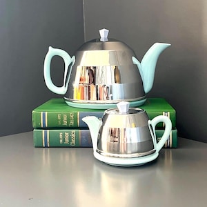 Op de afbeelding: Een grote en een kleine theepot, elk met een gepolijst chromen lichaam, een licht mintgroen handvat, tuit, voet en een witte geometrische knop op het deksel. De theepotten staan op twee groene boeken met de titel Junior Classics.