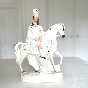 Puede incluir: Una figura de cerámica blanca que representa a un nativo americano a caballo. La figura viste ropa blanca y un tocado, con detalles en negro. El caballo es blanco con marcas negras.