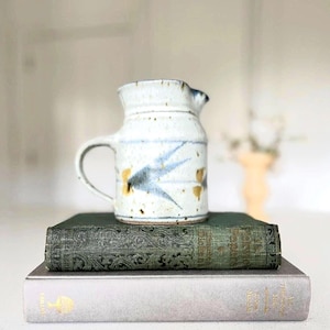 Op de afbeelding: Een kleine, gebroken witte keramische kan met een handvat en tuit, versierd met blauwe en gouden abstracte ontwerpen. De kan staat op een stapel van twee antieke boeken met groene en grijze covers.