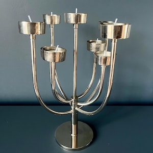 May include: A silver-toned metal candelabra featuring seven individual candle holders arranged at varying heights on curved arms. The structure is supported by a central vertical post and a circular base, with white candle wicks visible in each cup.