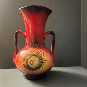 May include: A vintage ceramic vase in red and brown tones. The vase has two handles and a distinctive design on the body, with a yellow and orange centre and black lines radiating outwards. A decorative item for the home.