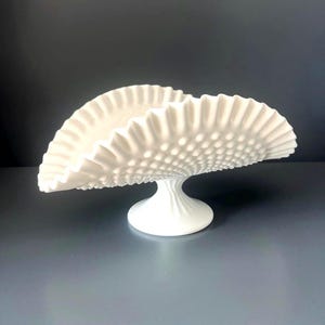 Puede incluir: Un cuenco de vidrio lechoso blanco con textura de puntos en relieve y un borde ancho, fruncido y ondulado. El cuenco se apoya sobre una base de pedestal estriada, creando una forma curva y alargada que se eleva hacia dos extremos.