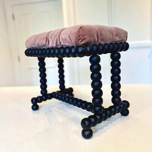 May include: A small, decorative stool with a plush, dusty rose cushion. The base is constructed of black, rounded wooden beads. The legs and frame are also black, creating a striking contrast with the cushion. The stool is designed for seating.