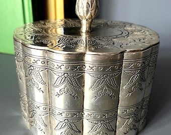 May include: A round silver-toned metal container with a lid featuring a textured artichoke-shaped finial. The exterior surface is decorated with repeating embossed floral patterns and horizontal bands, creating a scalloped edge profile.