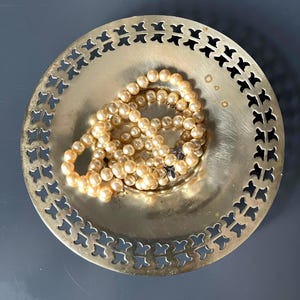 Peut inclure: Un plat circulaire en laiton avec un motif de bordure découpé contenant un collier de perles sphériques de couleur crème. La surface métallique présente des signes d'usure et d'oxydation, avec un éclat métallique et des perles au centre.