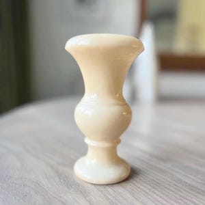 May include: A small, cream-colored decorative vase with a flared top and a rounded base. The vase has a classic, elegant shape, and is made of a smooth, glossy material. The vase is approximately 4 inches tall.
