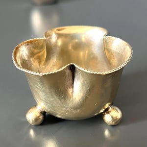 Può includere: Una piccola ciotola metallica color oro con un bordo smerlato e perlato, sostenuta da tre piedini sferici. La ciotola ha una superficie liscia e riflettente con segni visibili di usura e patina, caratterizzata da un'apertura ampia.