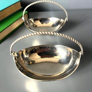 May include: Two silver-colored metal bowls with a polished, reflective finish. Each bowl features a semi-circular, twisted rope-style handle attached to the sides. The bowls have a smooth, curved interior and a wide, open top design.