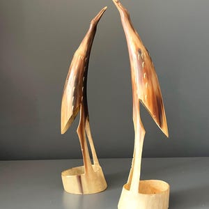 Può includere: Due sculture di uccelli alte e slanciate realizzate in corno levigato. Ogni figura presenta un lungo becco appuntito, un collo ricurvo e un corpo liscio e affusolato con sottili segni incisi simili a piume in tonalità crema, beige e marrone scuro.