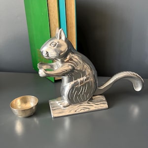 May include: A silver-toned metal nutcracker shaped like a squirrel standing on a rectangular base with a textured tail and a small, separate silver-toned metal bowl positioned to the left of the squirrel figure on a flat surface.
