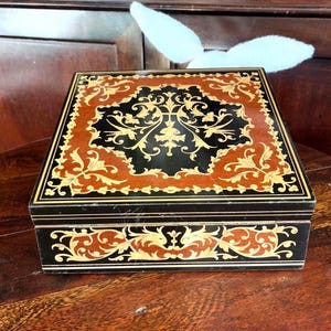 May include: A square wooden box with detailed inlaid patterns. The box has a dark brown base with a black and gold floral design on the lid. The sides are adorned with gold and brown scrollwork. A decorative storage item.