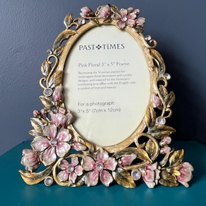 May include: An oval-shaped gold-toned picture frame featuring ornate floral embellishments in shades of pink and green. The frame includes clear crystal accents and text reading Past Times, Pink Floral 3" x 5" Frame, and For a photograph 3" x 5".