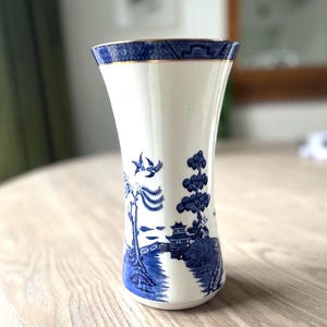 May include: A white porcelain vase with a blue and gold rim and a blue landscape design. The vase features a detailed illustration of a bridge, trees, and birds. The vase is sitting on a wooden table.