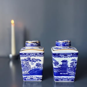 以下が含まれることがあります： 青と白の伝統的なウィローパターンが施された2つの四角い陶器製ジャー。各ジャーには金色の縁取りが施された蓋が付いています。デザインには、青いインクで描かれた塔、橋、木々などの伝統的な風景が表現されています。