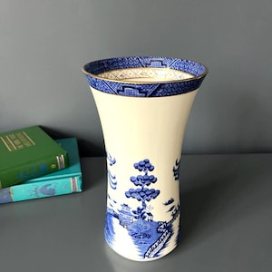 May include: A cream-coloured ceramic vase featuring a blue willow pattern with trees, buildings, and bridges. The flared rim is decorated with a blue and white geometric border and a thin gold trim along the top edge of the cylindrical vessel.