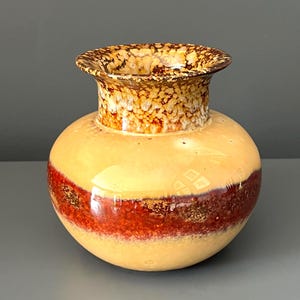 Puede incluir: Un jarrón de cerámica con cuerpo redondo y cuello acampanado. El jarrón presenta una paleta de colores cálidos, con una base amarillo claro, una ancha banda marrón rojizo y un borde marrón moteado. El acabado brillante realza los colores.