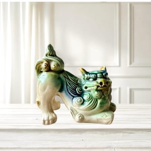 May include: A ceramic, stylized lion-dog figurine in shades of green, blue, and cream. The mythical creature has detailed features, including curled mane and tail, and is posed in a standing position. The figurine is on a white surface.
