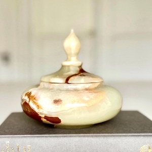 May include: A decorative, lidded jar made of onyx. The jar has a bulbous body and a pointed finial on the lid. The onyx is a pale green color with brown and cream-colored veining. The jar is sitting on a gray book.