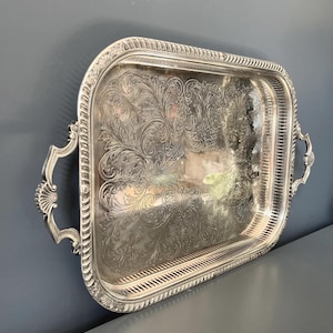 Puede incluir: Una bandeja de servicio rectangular de tono plateado que presenta un intrincado patrón floral grabado en la superficie central. La bandeja tiene un borde elevado con una orilla acanalada y dos asas curvas ornamentadas en los lados con detalles de concha.