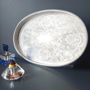 Op de afbeelding: Een ovaal zilverkleurig dienblad met een ingewikkeld reliëfpatroon van bloemen en krullen, voorzien van een touwachtige rand. Daarnaast staan twee glazen parfumflesjes, één met een blauwe dop en één met een rood- en goudkleurige conische dop.