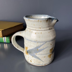Op de afbeelding: Een keramieken kan met een gespikkeld gebroken wit glazuur, voorzien van een blauw en bruin geschilderd bladachtig patroon rondom het lichaam. De kan heeft een gebogen handvat aan de zijkant, een uitlopende rand en een kleine schenktuit.